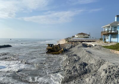 A bulldozer works in the surf to carefully move material against existing residential seawalls